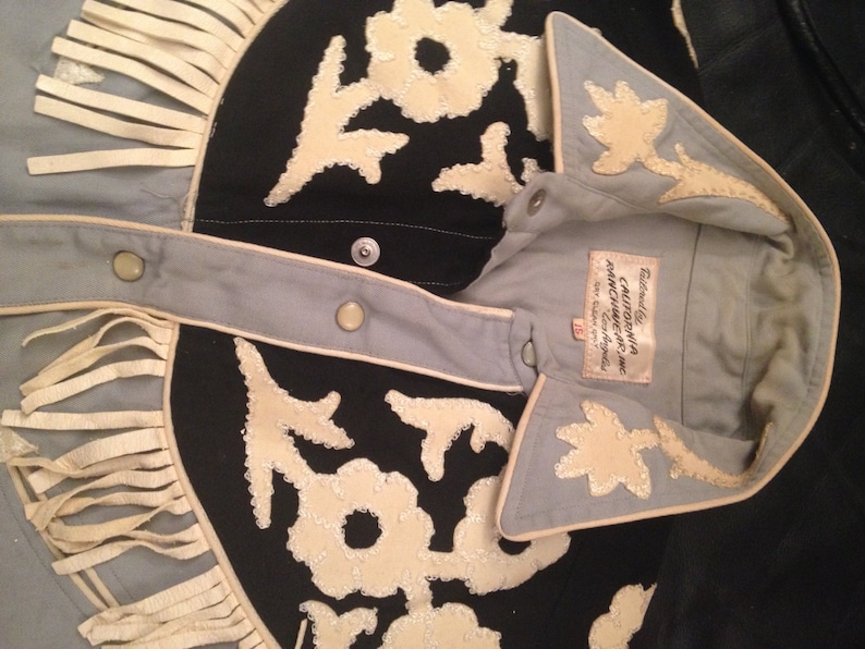 May include: A light blue denim shirt with a black and white embroidered design. The shirt has a white fringe trim and a white collar. The label reads "California Ranchwear Inc. Los Angeles."