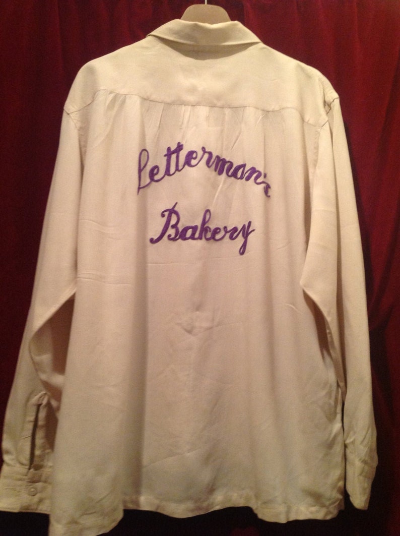 May include: A white long-sleeved shirt with a purple embroidered logo that says "Letterman's Bakery".