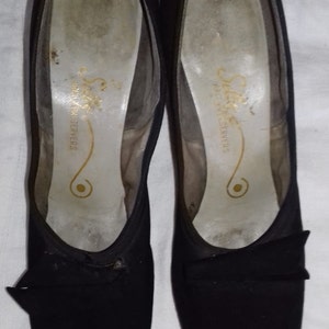 May include: A pair of black peep-toe heels with a bow detail. The shoes are made of a soft, suede-like material and have a simple, elegant design. The inside of the shoes has the text "Selma Fashion Servers" printed on the insole.