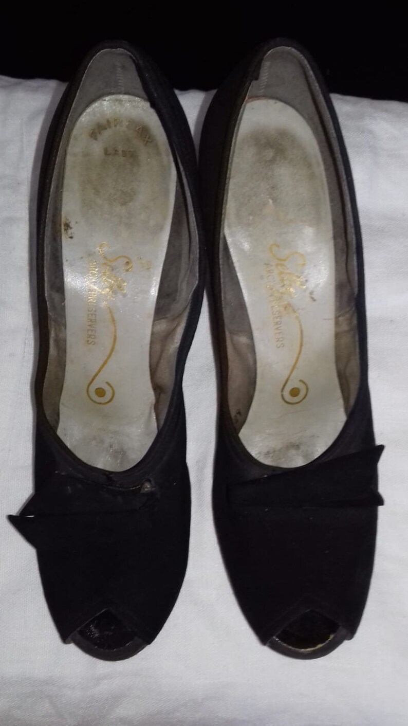 May include: A pair of black peep-toe heels with a bow detail. The shoes are made of a soft, suede-like material and have a simple, elegant design. The inside of the shoes has the text "Selma Fashion Servers" printed on the insole.