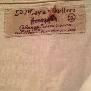 May include: A tan fabric label with brown text that reads "La Play'a by Bariboro", "Celanese", "FABRIC OF RAYON", "REG. U.S. PAT. OFF.", "HAND WASHABLE - PRESS WITH COOL IRON", and "Honey Sun". The label also has a size tag that reads "XL".