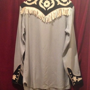 May include: A light blue long-sleeved shirt with black and white embroidery on the yoke and cuffs. The yoke has a fringe trim.