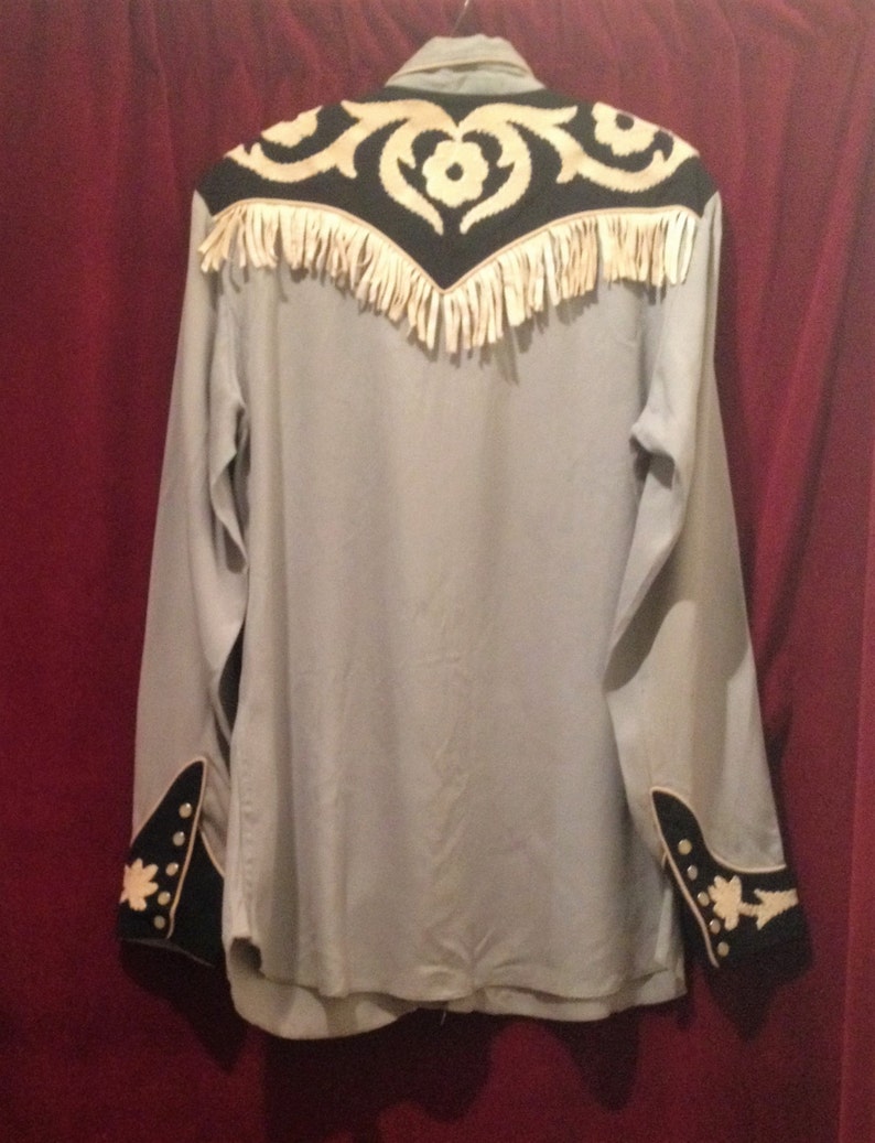 May include: A light blue long-sleeved shirt with black and white embroidery on the yoke and cuffs. The yoke has a fringe trim.
