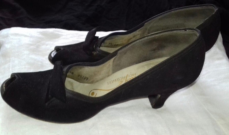 May include: A pair of black peep-toe heels with a low heel. The shoes are made of a soft, suede-like material and have a simple, elegant design.