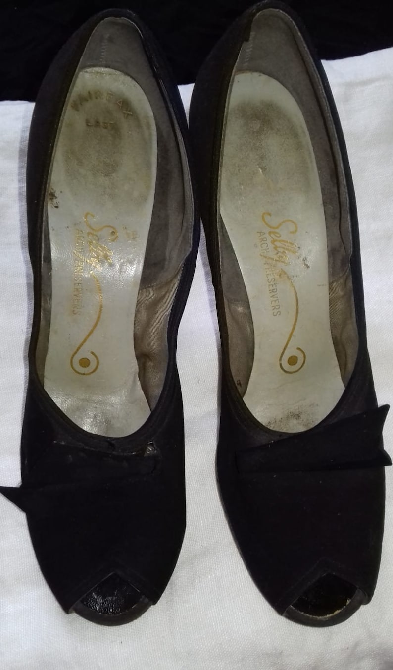 May include: A pair of black peep-toe heels with a bow detail. The shoes are made by Selkirk Arch Preservers.