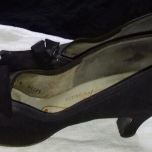 May include: A pair of black peep-toe heels with a small bow detail on the front. The shoes have a low heel and are made of a soft, suede-like material.