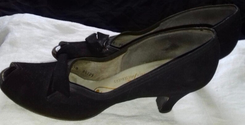 May include: A pair of black peep-toe heels with a small bow detail on the front. The shoes have a low heel and are made of a soft, suede-like material.