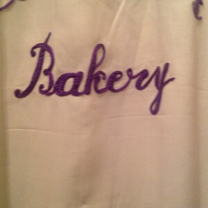 May include: A white fabric sign with purple embroidery that reads "Bellemann's Bakery".