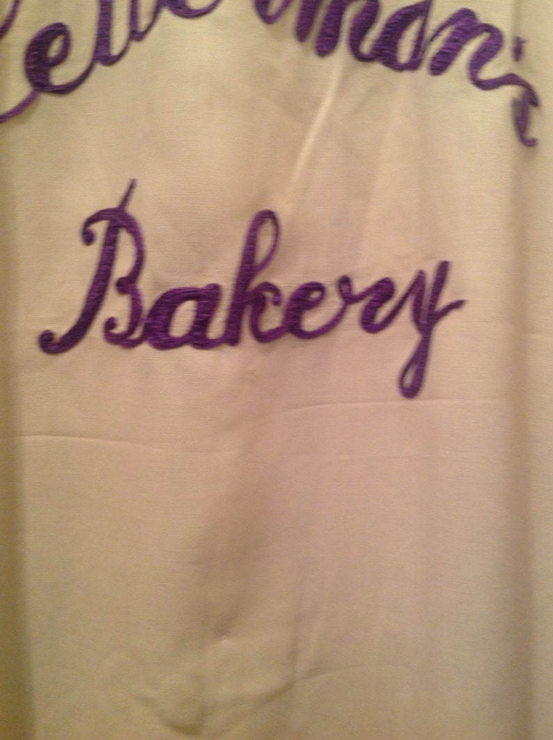 May include: A white fabric sign with purple embroidery that reads "Bellemann's Bakery".