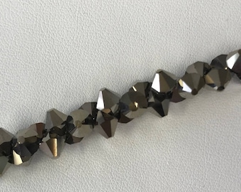 Può includere: Un filo di perline sfaccettate grigio scuro. Le perline hanno una forma geometrica a più lati e sono infilate su un filo sottile e trasparente. Le perline sono disposte in fila su uno sfondo bianco.