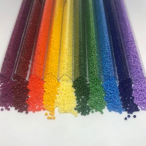 May include: Assortment of colorful seed beads in clear plastic tubes. The beads are arranged in a rainbow pattern, starting with burgundy and ending with lavender.