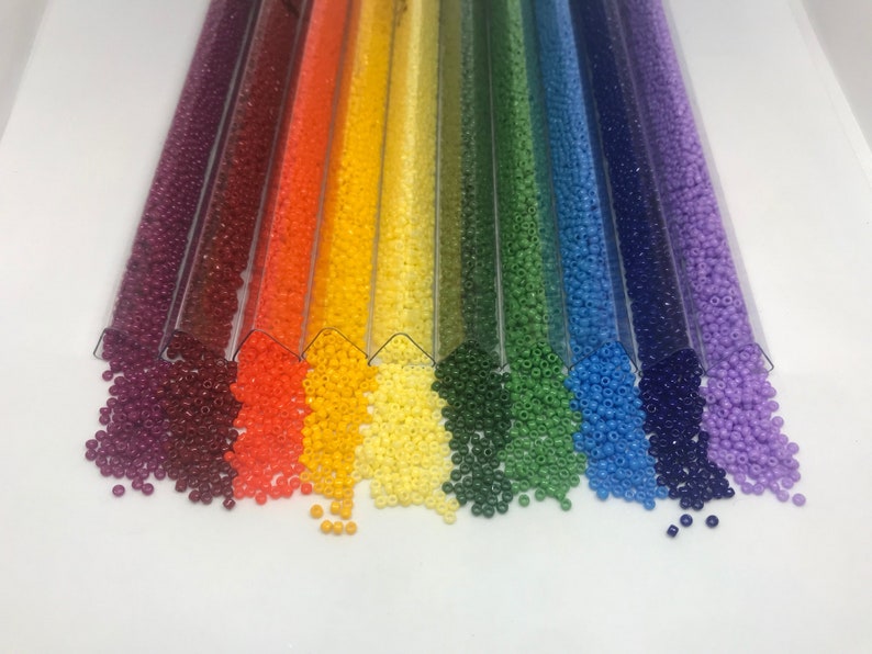 May include: Assortment of colorful seed beads in clear plastic tubes. The beads are arranged in a rainbow pattern, starting with burgundy and ending with lavender.
