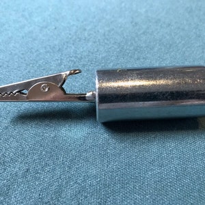 May include: A silver-colored alligator clip with a cylindrical metal body. The clip has a serrated jaw and is attached to the body. The object is set against a teal-colored fabric background.