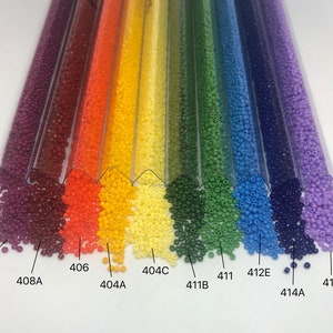 May include: A rainbow assortment of seed beads in various colors, including purple, red, orange, yellow, green, blue, and light purple. The beads are arranged in rows in clear plastic tubes, with the color of the beads spilling out of the tubes. The tubes are labeled with numbers, including 408B, 408A, 406, 404A, 404C, 411B, 411, 412E, 414A, and 419.