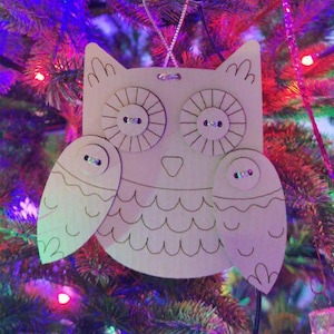 May include: A wooden owl Christmas ornament with button eyes and wing details, hanging on a decorated Christmas tree. The owl is made of light-colored wood and has a simple, outlined design. The background is a blur of colorful Christmas lights.