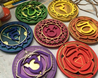 May include: Seven round wooden ornaments painted in the colors of the seven chakras. Each ornament features a stylized symbol and the Sanskrit word for the chakra. The colors are red, orange, yellow, green, blue, indigo, and violet.