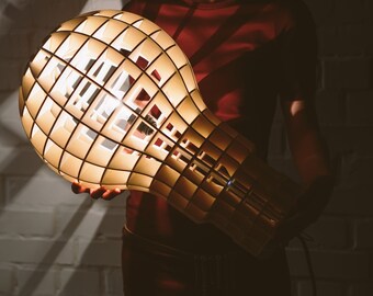 May include: A large, wooden light bulb shaped lamp with a grid pattern. The lamp is lit and casting a warm glow.