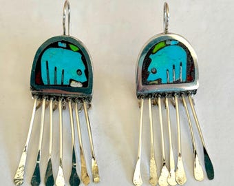 May include: A pair of silver dangle earrings featuring a semi-circular inlay of bright turquoise stone shaped like a bear, accented with small green and red stone details. Each earring has five thin, flat silver fringe strands hanging below.