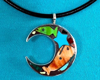 May include: A silver crescent moon pendant necklace with a black leather cord. The pendant is inlaid with turquoise, red, green, and brown stones, and features a star and two fish designs.