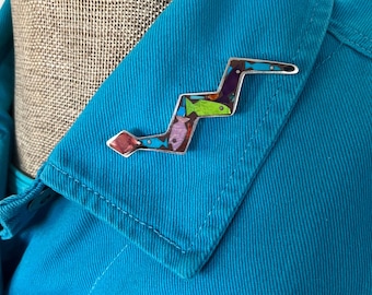 May include: A silver brooch in the shape of a zig-zag line with colorful fish designs. The brooch is on a blue fabric background.
