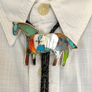 May include: A bolo tie featuring a horse-shaped pendant crafted with inlaid multicolored stones including turquoise, mother-of-pearl, and coral. The pendant is attached to a black braided leather cord and rests against a white button-down shirt.