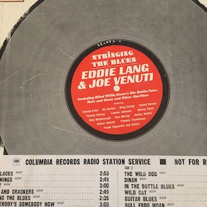 May include: A black and white record label with the text "Stringing the Blues Eddie Lang & Joe Venuti" and a list of songs on the record. The record label is for Columbia Records Radio Station Service.