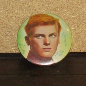 May include: A vintage button featuring a color portrait of a young man with red hair. The button has a green background with the name "Dale" written in cursive on the bottom right and "Hawley" written in cursive on the top right.