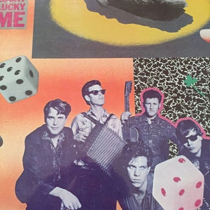 May include: Album cover for The Leroi Brothers' "Lucky Lucky Me." The image features a band of five men in vintage attire, with dice and a lucky charm graphic. The background is a mix of orange, black, and white patterns.
