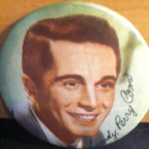 May include: A black and white photo of a man with dark hair. He is wearing a suit and smiling. The photo is signed "Chris Perry".
