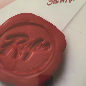 May include: A close-up of a red wax seal with the name "Rufus" and the text "Seal in Red" on a white background. The seal is circular with a raised design. The image is well-lit and the colors are vibrant.