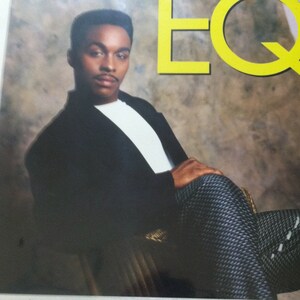 May include: A black and white photo of a man wearing a black suit jacket, a white shirt, and black and white patterned pants. He is sitting in a chair and looking at the camera. The word "EQ" is written in yellow letters in the upper right corner of the image.