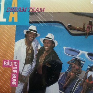 May include: A record album cover featuring two men wearing white hats and gold chains. The album cover is titled "L.A. Dream Team" and the album title is "Bad to the Bone". The background features a swimming pool and people relaxing by the pool.