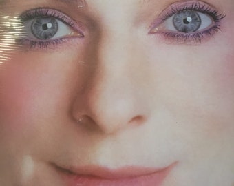 May include: Close-up of a woman's face with soft, natural makeup. Her eyes are highlighted with light purple eyeshadow and her lips are a soft pink. The image is slightly blurred, giving it a dreamy, ethereal quality.