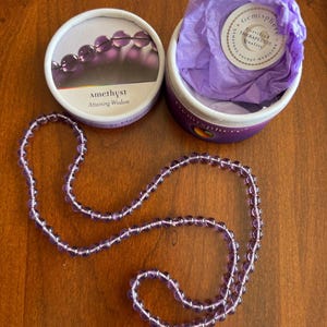 May include: A strand of round, translucent purple amethyst beads arranged in a loop. Beside it sits a white cylindrical container with a lid featuring an image of amethyst beads and the text Amethyst Attaining Wisdom, and a base containing purple tissue paper.