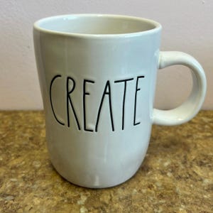 May include: White ceramic mug with the word "CREATE" in black, handwritten-style lettering. The mug has a rounded shape and a curved handle. The mug is sitting on a brown surface.