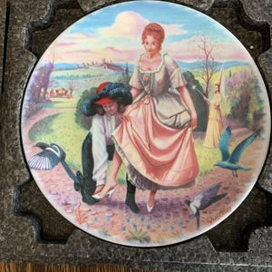 May include: A round decorative porcelain plate featuring a colorful illustration of a person in a long peach-colored dress and a person in a white shirt and black pants. The scene includes birds, a distant castle, and the signature Quellier.