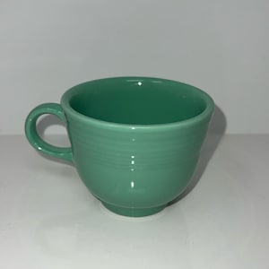 Puede incluir: Una taza de té de cerámica verde pastel con un asa curva y un cuerpo redondeado. La taza presenta estrías horizontales en el exterior y una pequeña base circular. El color es un verde suave y apagado.