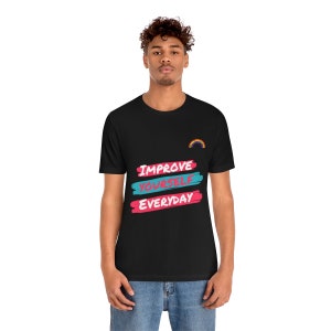 Op de afbeelding: Zwart T-shirt met de tekst "IMPROVE YOURSELF EVERYDAY" in roze en blauwe verfstreken. Een kleine regenbooggrafiek bevindt zich in de rechterbovenhoek.
