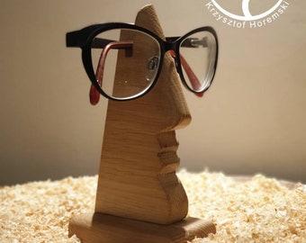 Puede incluir: Portagafas de madera con forma de cara con un par de gafas negras apoyadas en la nariz. El soporte está sobre un lecho de virutas de madera.