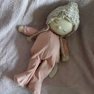 May include: A pink fabric doll with a crocheted hat, lying on a white towel. The doll has a simple face with closed eyes and a stitched smile.