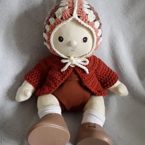May include: A crocheted doll wearing a brown, pink and white crocheted hat, a red crocheted cardigan, and brown shoes.