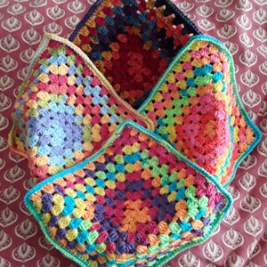 May include: Four colourful, square crochet pieces stacked together. Each piece has a granny square design with vibrant pink, orange, yellow, green, blue, and purple hues. The edges are finished with a contrasting border.