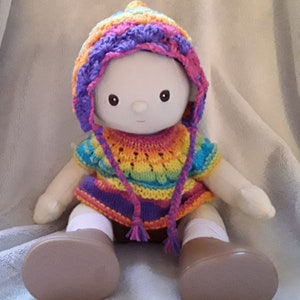 May include: A white doll wearing a rainbow crocheted hat and matching sweater. The doll has brown eyes and a sweet expression.