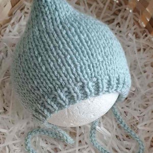 May include: A pale blue knitted baby bonnet with ear flaps and ties. The bonnet is made of a soft, textured yarn and is displayed on a white spherical form. The background includes a woven basket and white paper.