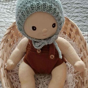 May include: A handcrafted fabric doll, wearing a pale blue knitted hat and scarf, and a brown romper. The doll has a neutral skin tone and sits in a woven basket with paper. The doll has dark eyes and a simple facial expression.