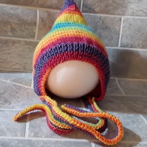 May include: A rainbow striped knitted hat with a pointed crown and long ties. The hat is made with a variety of colours, including red, orange, yellow, green, blue, and purple.