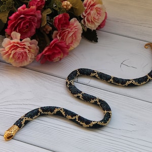 Op de afbeelding: Een kralen slangenketting met een zwart en gouden patroon. De ketting ligt op een wit houten oppervlak.