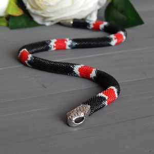 May include: A beaded necklace in the shape of a snake with black, red, and white beads. The snake has a silver head.