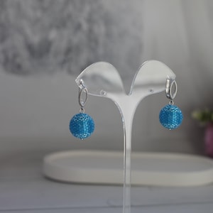 Può includere: Un paio di orecchini pendenti con un design a sfera blu chiaro con perline. Gli orecchini hanno ganci argentati a forma ovale. Sono esposti su un supporto in acrilico trasparente, con sfondo sfocato e fiori rosa.