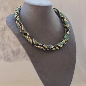 May include: A beaded necklace with a geometric pattern in black, green, and gold. The necklace is displayed on a gray bust. The beads are tightly woven, creating a textured surface. The design is a repeating pattern of angular shapes.